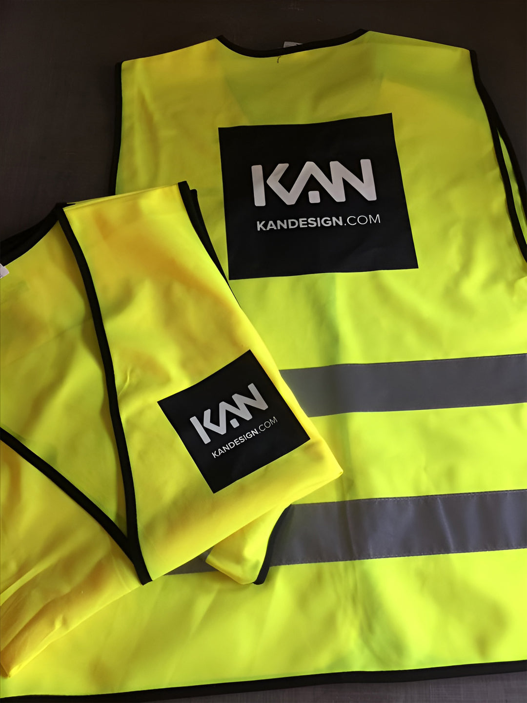 Two yellow safety vests with a black logo and website text on a dark surface.