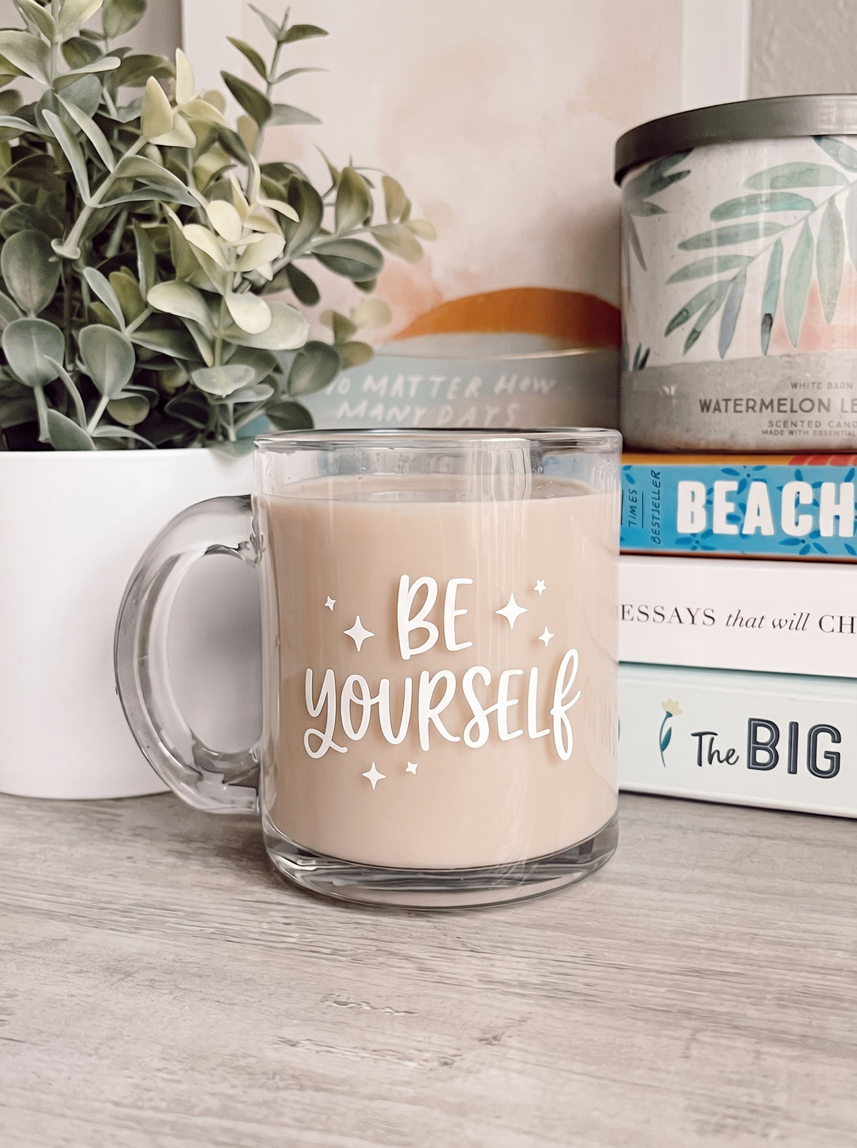 A clear glass mug filled with a drink sits on a wooden surface, customized with the phrase “BE YOURSELF” in white script and small star accents by Print365 Kenya