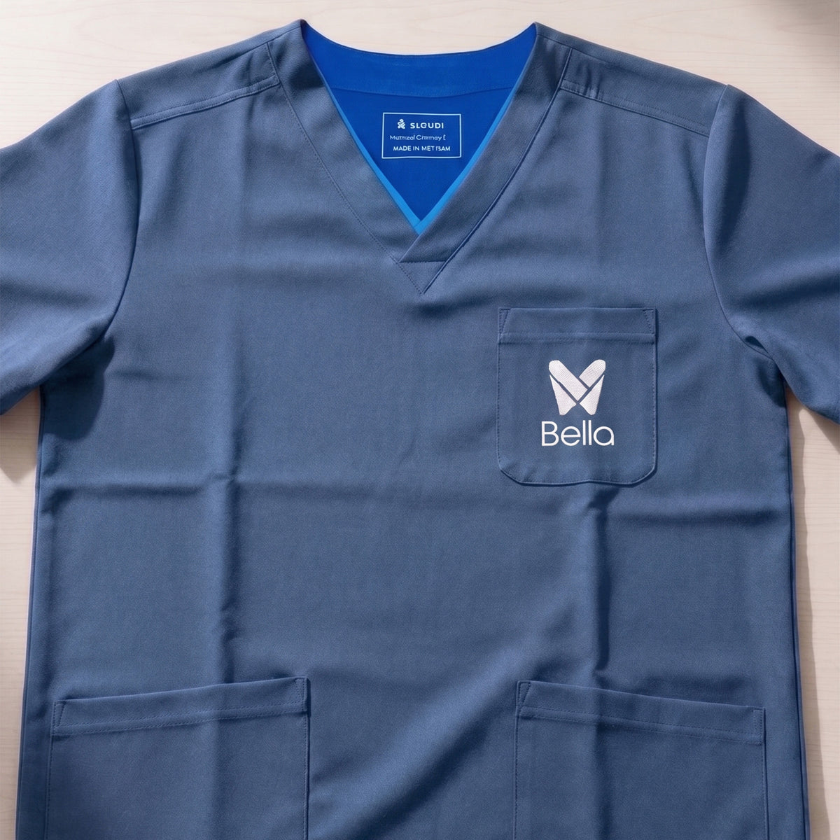 Custom slate blue scrub top with a V-neck, short sleeves, and two front pockets, featuring a white embroidered logo on the left chest pocket by Print365 Nairobi